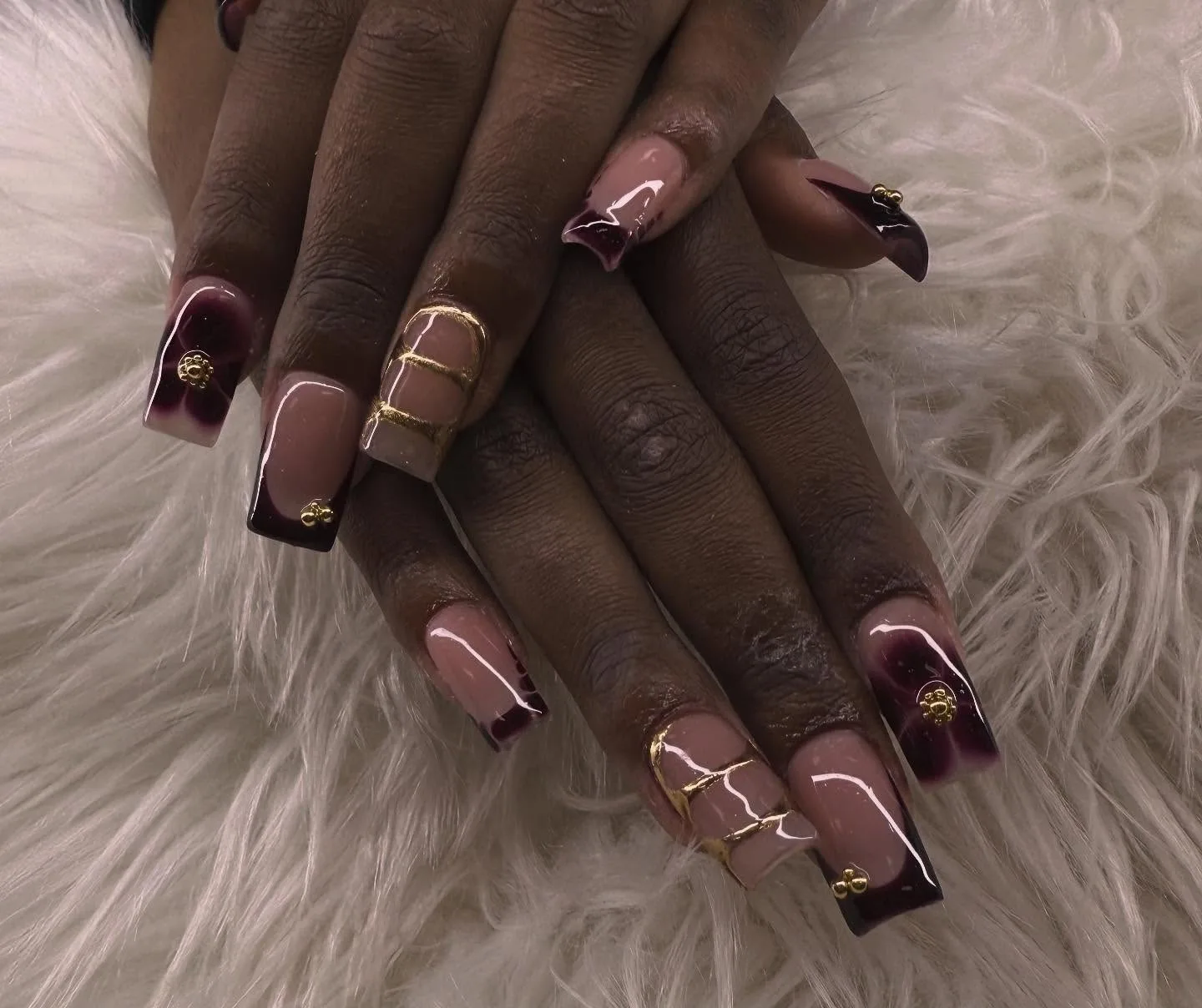 Long square nails on deep-brown interlaced hands — plum-to-clear ombré tips with gold beaded charms and gold wire wraps, glossy — on white fur
