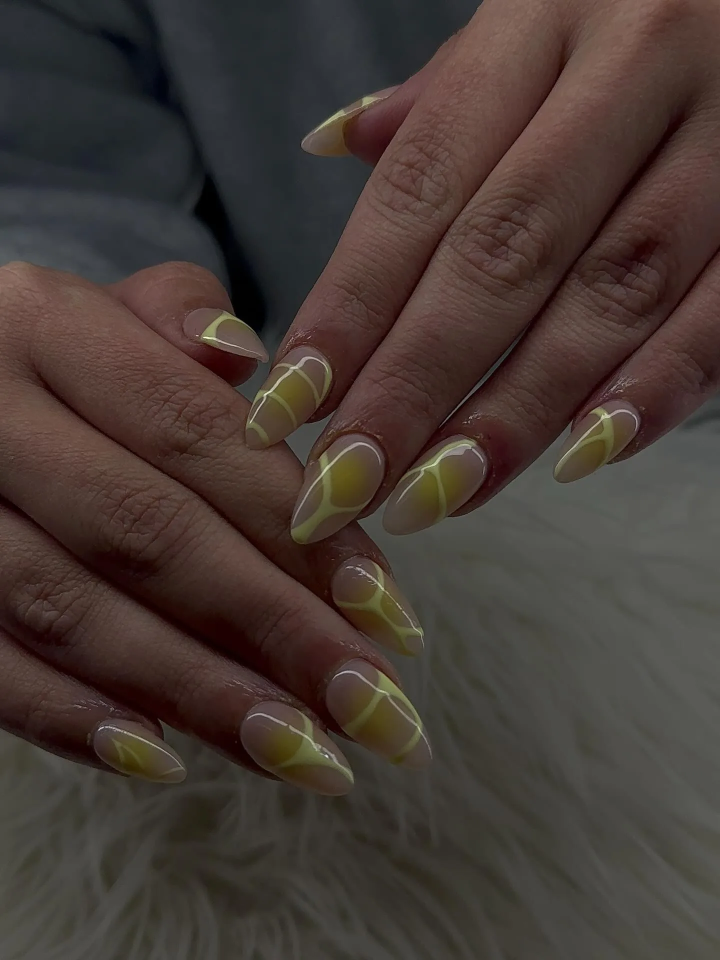 Almond nails with soft chartreuse-yellow swirl marbling over a nude base, two hands interlaced, cool muted light