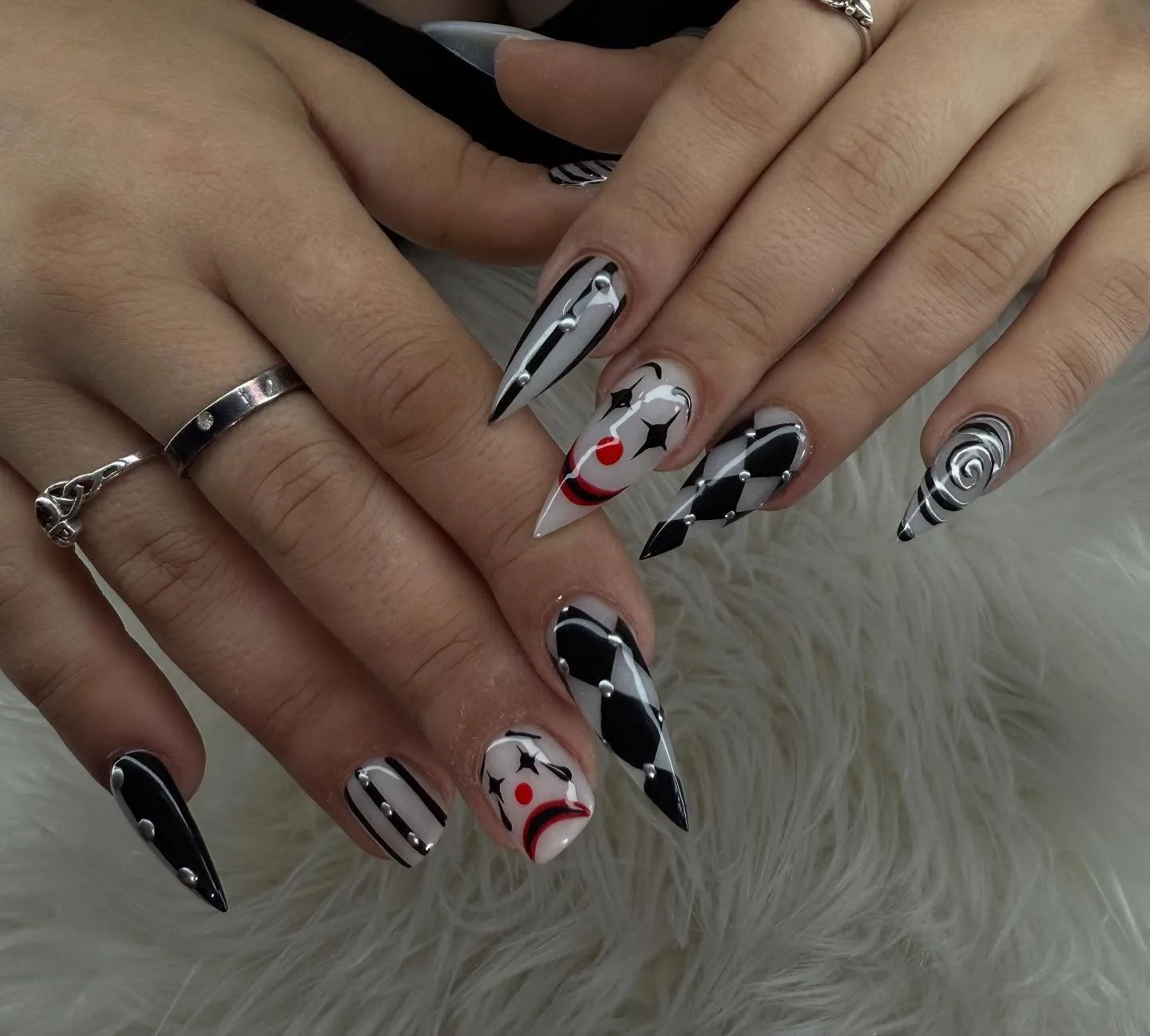 Long stiletto monochrome harlequin set — black-and-white diamonds, stripes, spirals and freehand-painted clown faces, chunky silver rings — on white fur