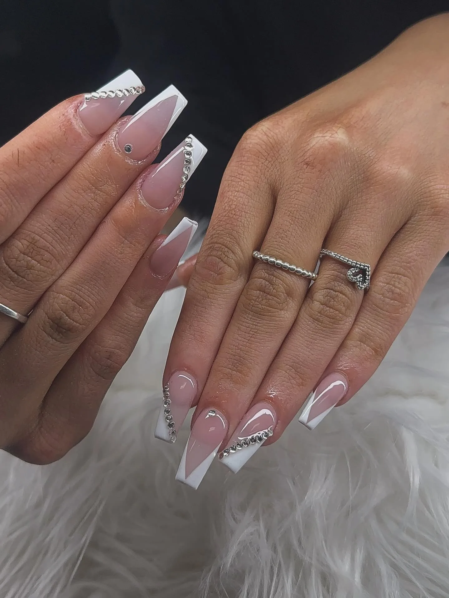 Long coffin and stiletto nude nails with white V-French tips and clear rhinestone strips along the smile line, beaded and heart silver rings — on white fur
