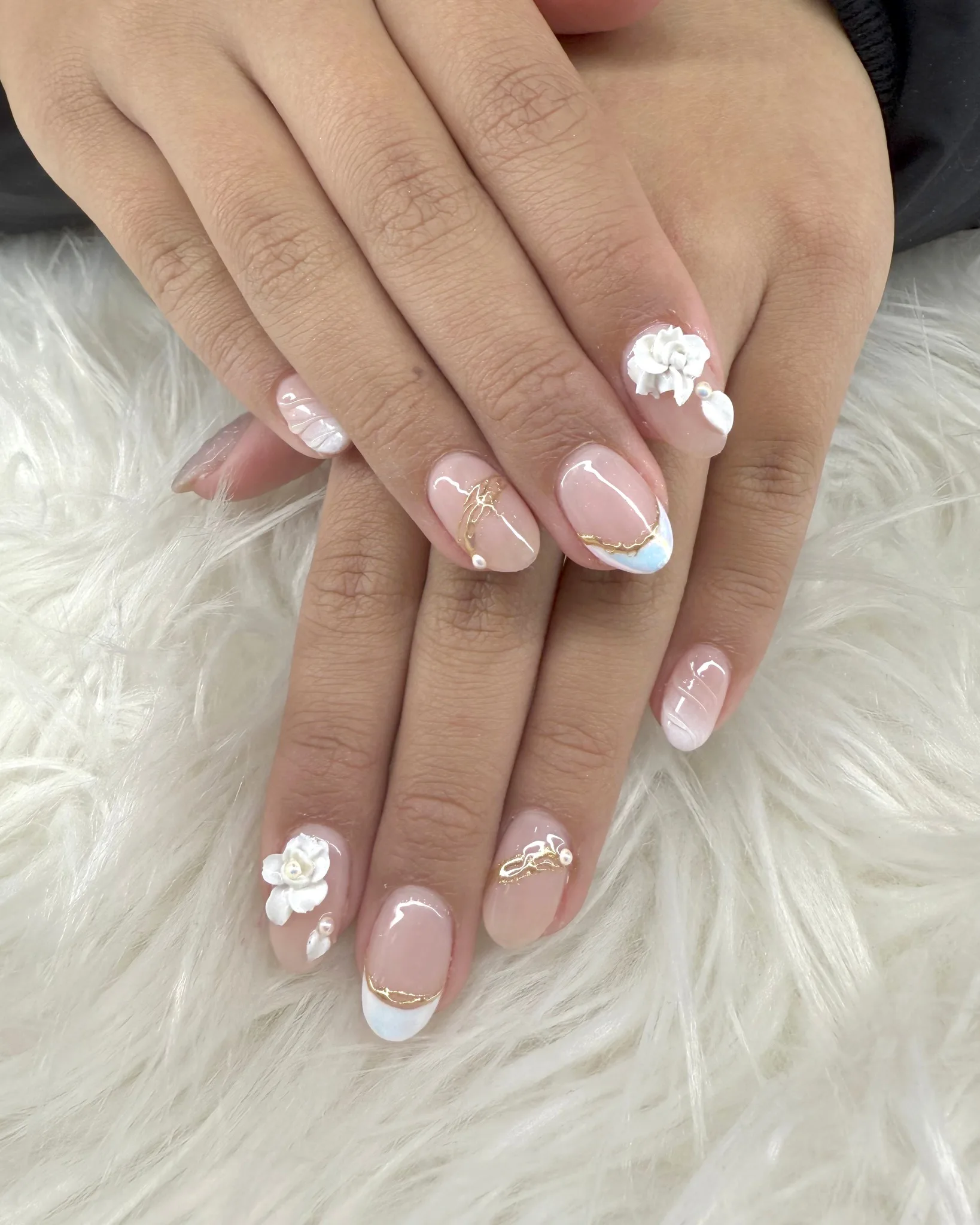Short round nude nails with hand-sculpted white 3D roses, real pearls, gold-foil veining and pale blue-gold French edge — on white fur