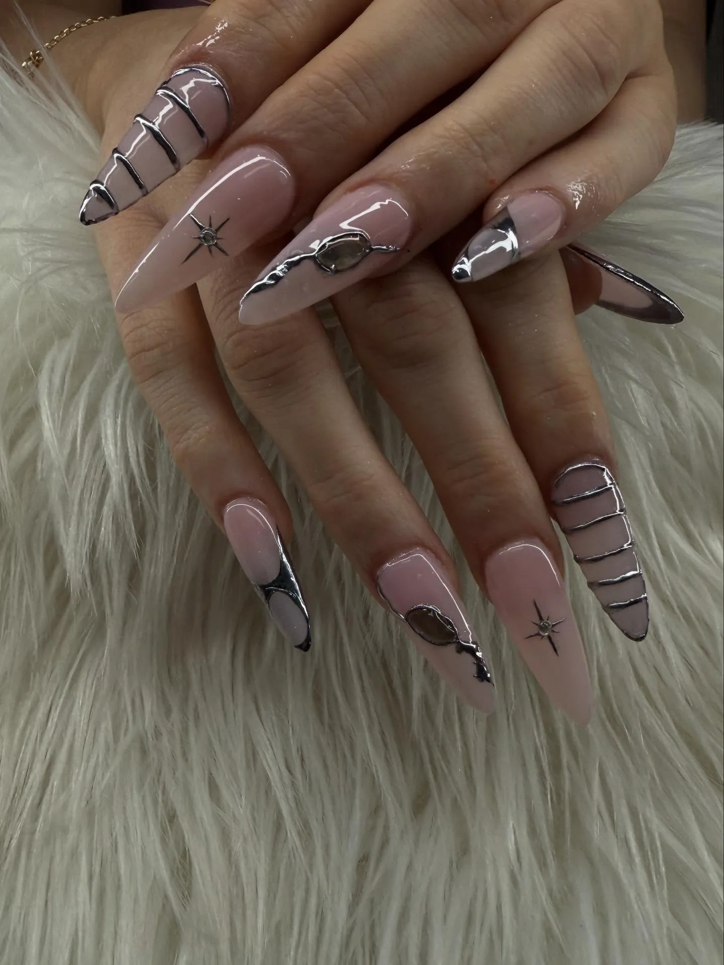 Long stiletto nude nails with liquid-chrome silver 3D drips, smoky gem inlays and engraved starburst accents, gold chain bracelet — on white fur