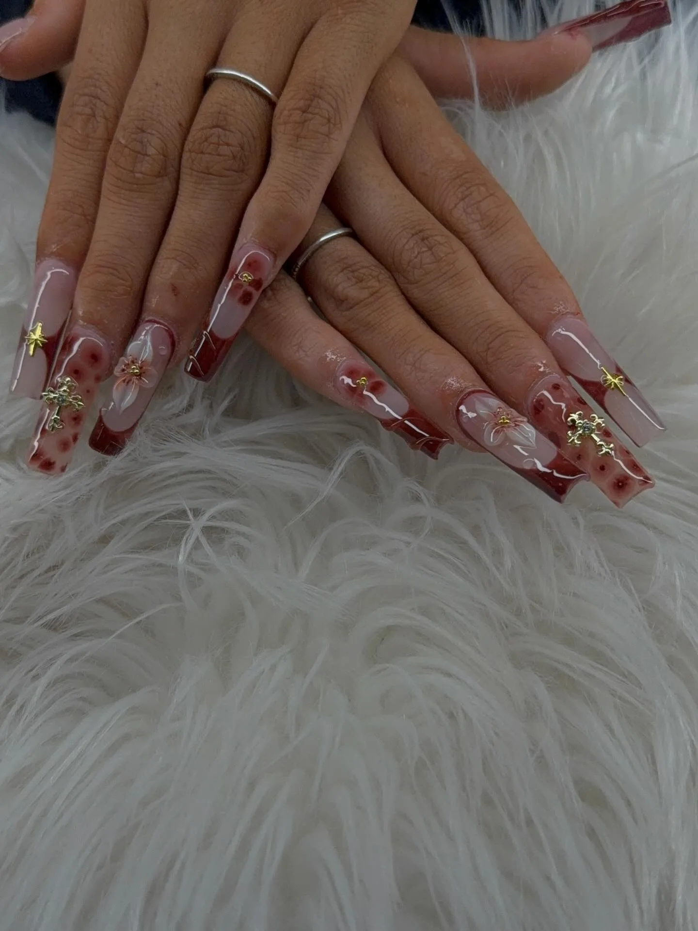 Extra-long coffin nails in deep rust-red and burgundy with clear floral inlays, red speckle art, gold cross and star charms, silver rings — on white fur