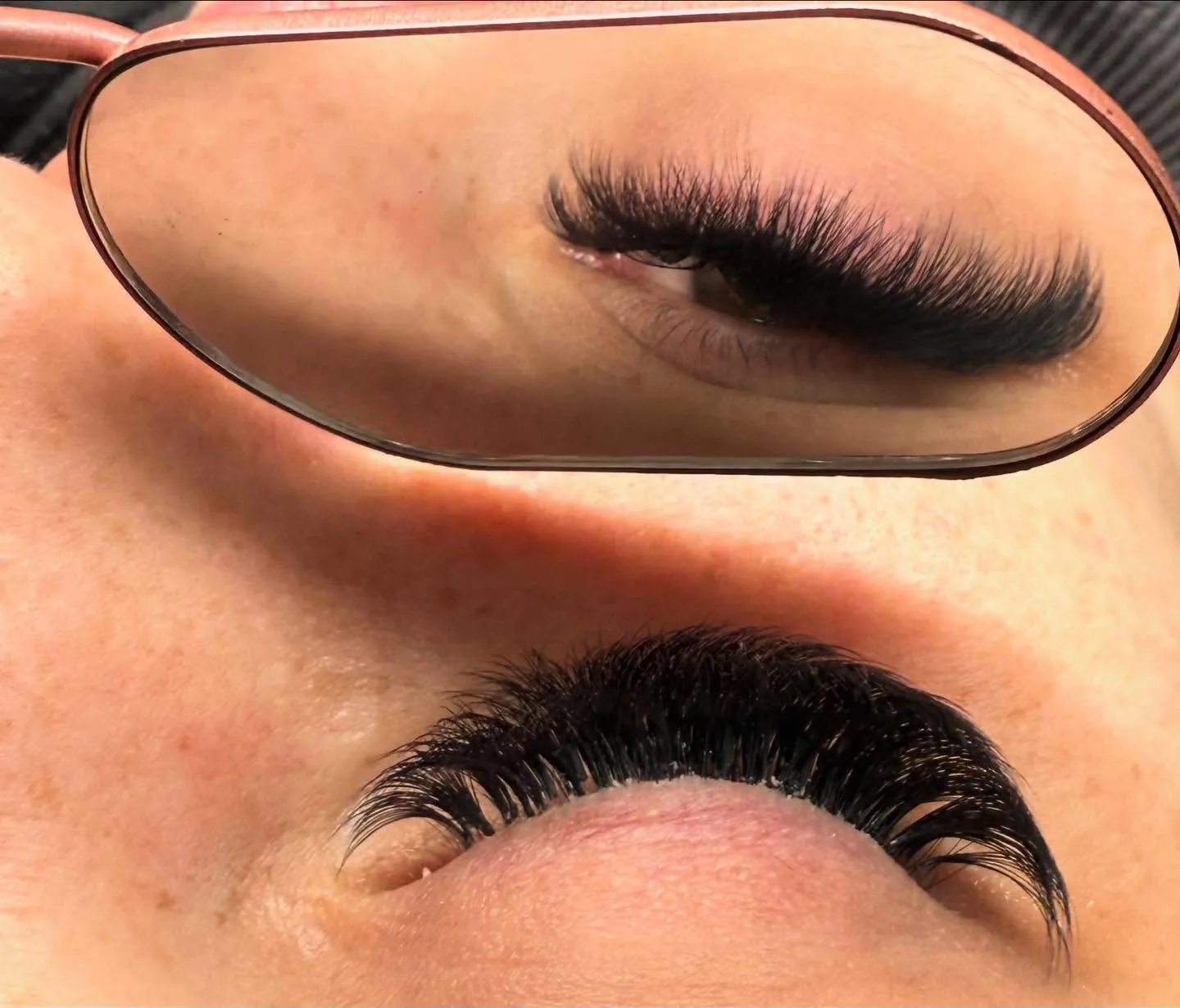 Close-up of a client's closed eye with a dense black lash set, a hand mirror angled above reflecting the same lash line