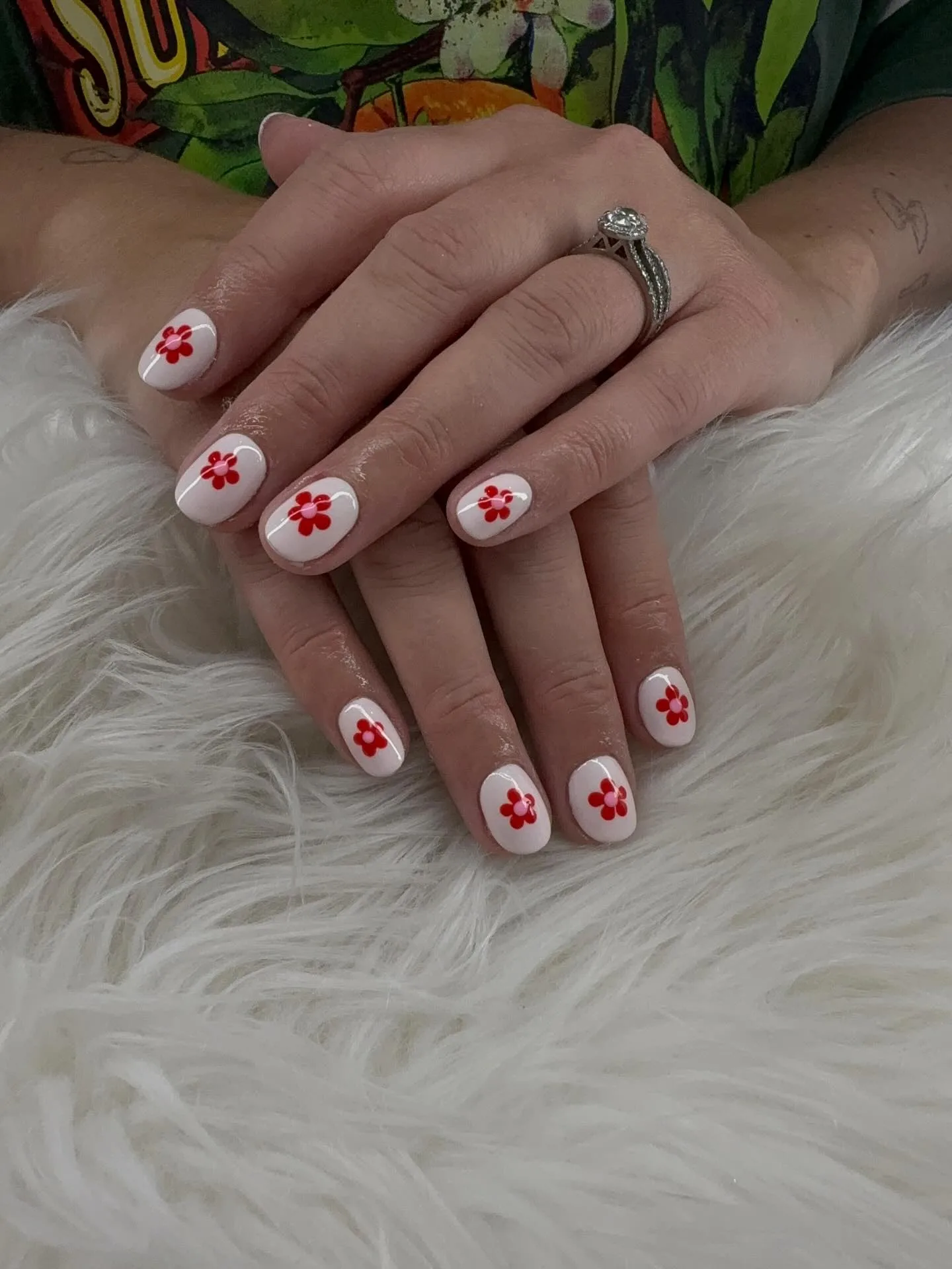 Short round milky-white nails with red-and-pink daisy motifs, diamond engagement ring, tropical-print sleeve — on white fur