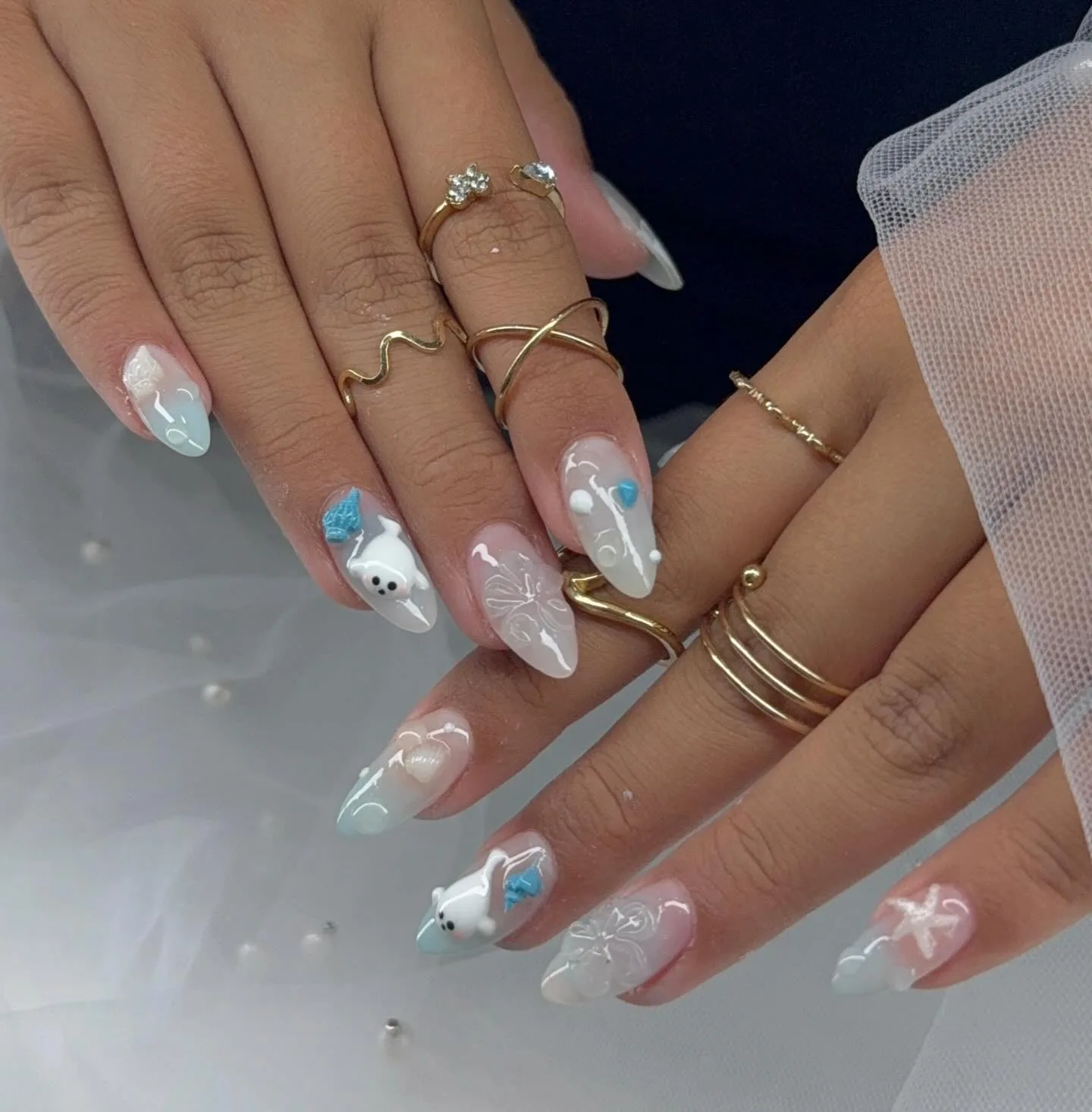 Almond nails in milky white and pale blue with sculpted 3D ghost-bear characters holding blue bows, white butterfly line art, stacked fine gold rings — on tulle with scattered pearls